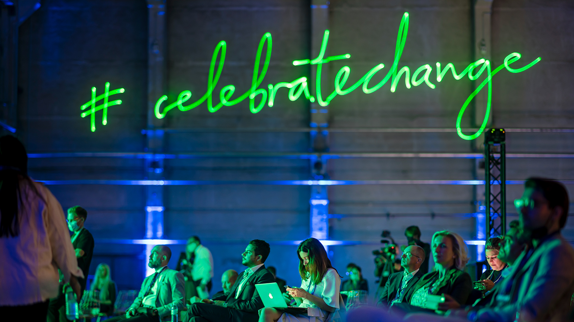 The green “#celebratechange” hashtag shines above the heads of audience members. 