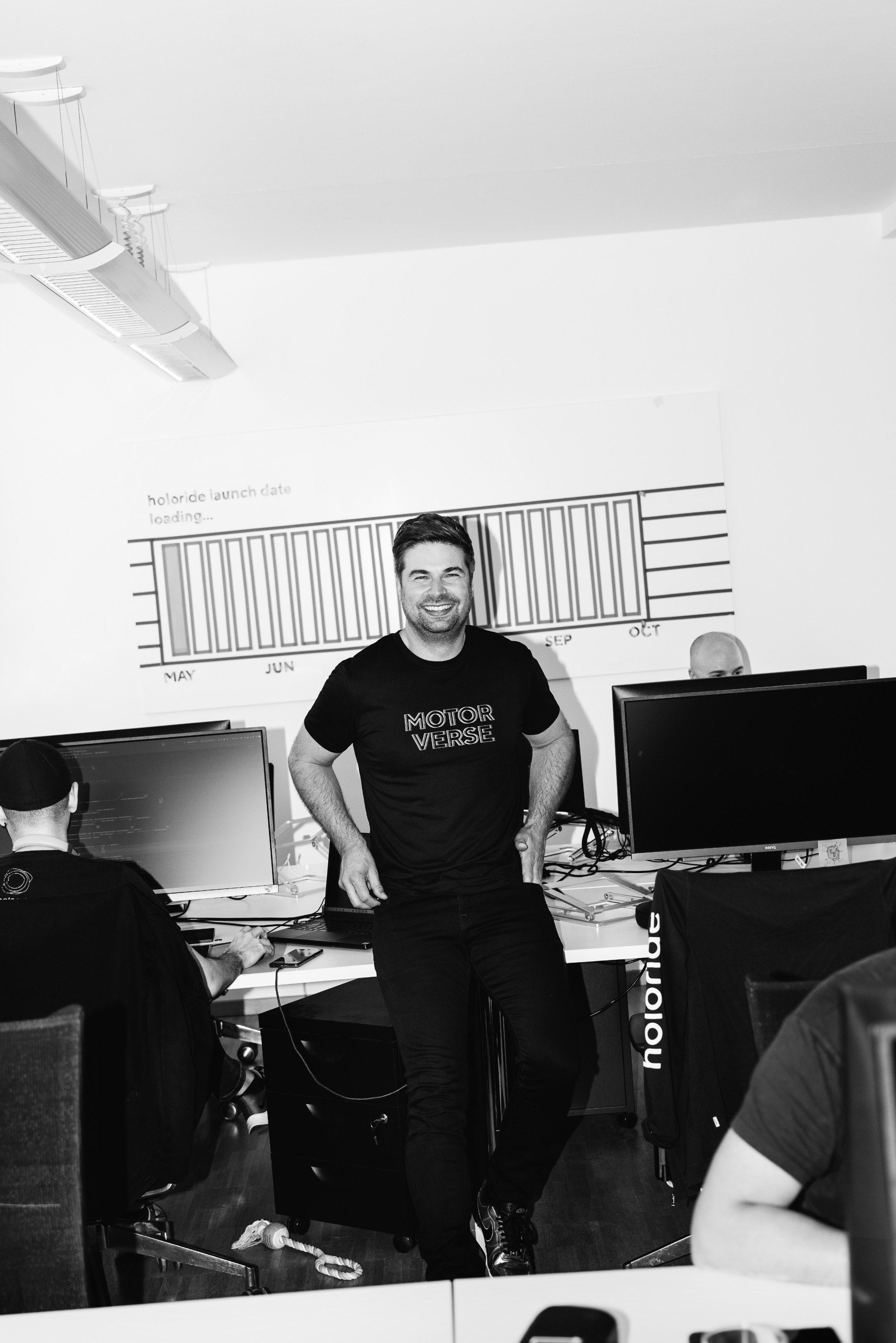 Nils Wollny standing at his desk surrounded by his team. His T-shirt reads "Motor Verse".